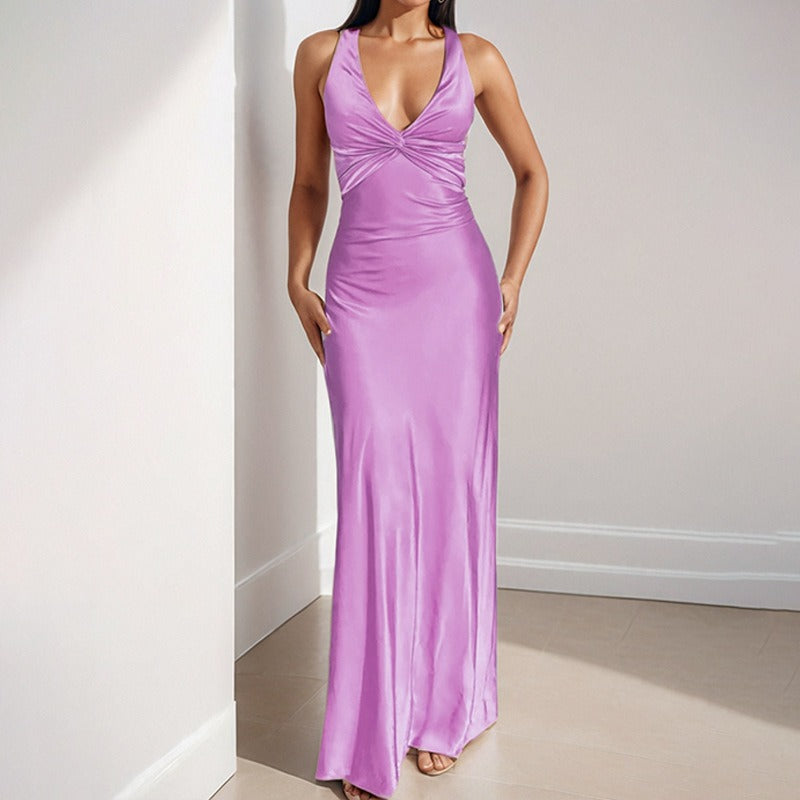 Woman wearing a lavender satin maxi dress with a twisted V-neckline and halter straps, standing in a corner.