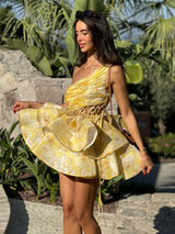 Woman wearing a yellow floral print one-shoulder mini dress with a ruffled tiered skirt and lace-up waist detail, posing outdoors.