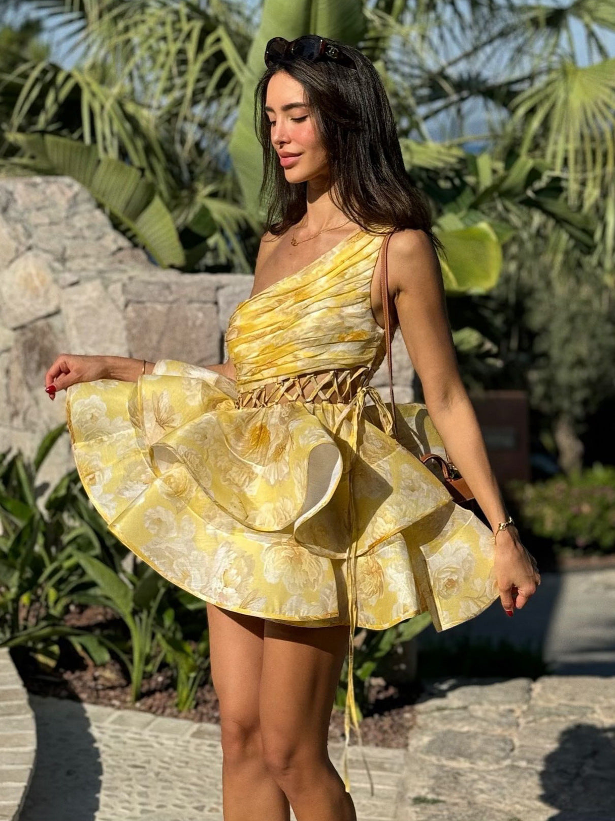 Woman wearing a yellow floral print one-shoulder mini dress with a tiered ruffled skirt, posing outdoors.