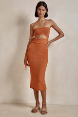 Woman wearing an orange strapless knit crop top and matching midi skirt with a tie waist and side slit.