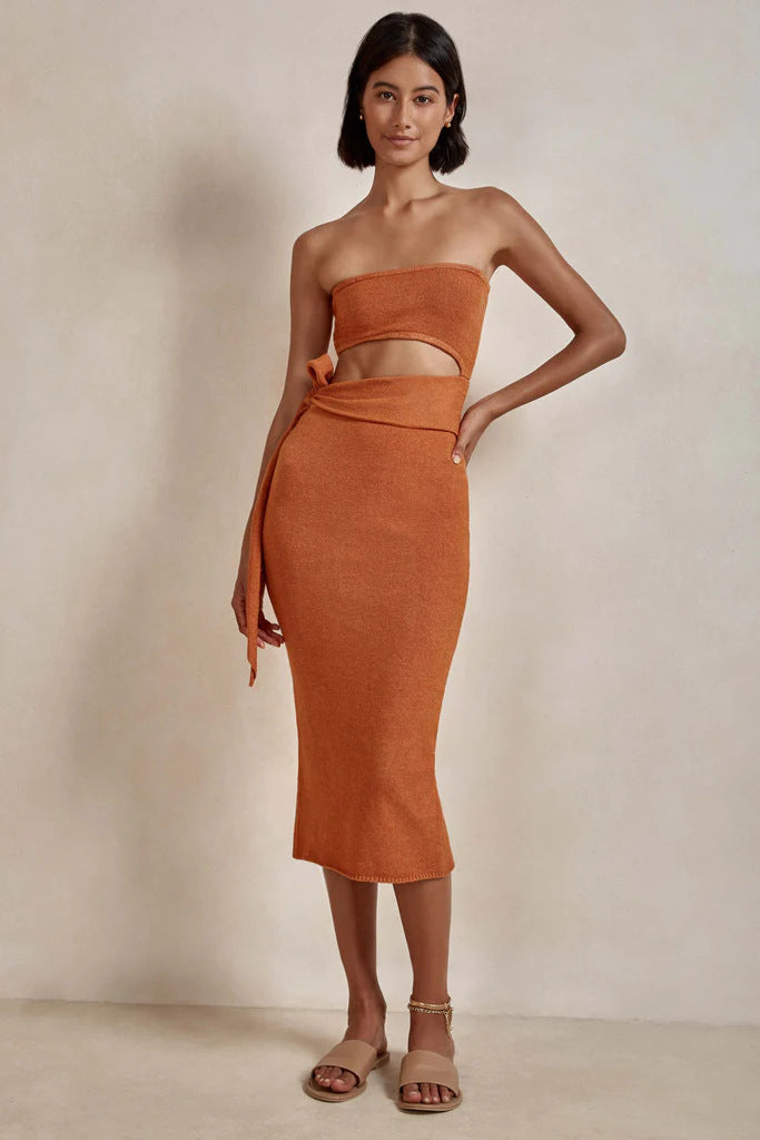 Woman wearing an orange strapless knit crop top and matching midi skirt with a tie waist and side slit.