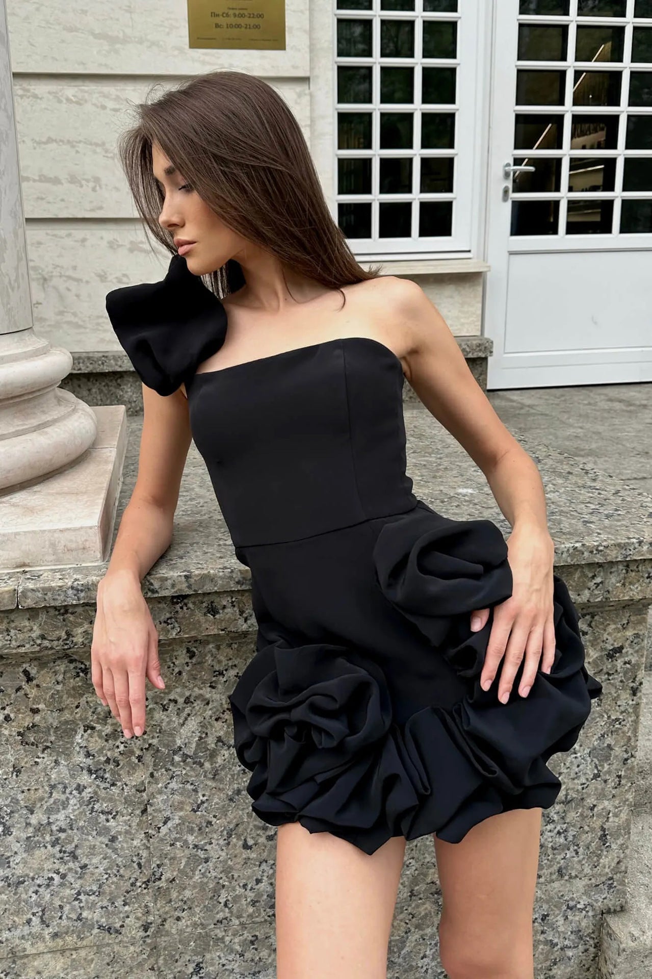 Woman leaning against a stone ledge wearing a black off-the-shoulder mini dress with ruffle details