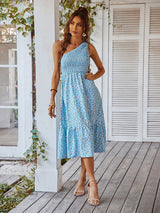 Woman wearing a light blue floral print one-shoulder midi dress standing on a wooden deck.