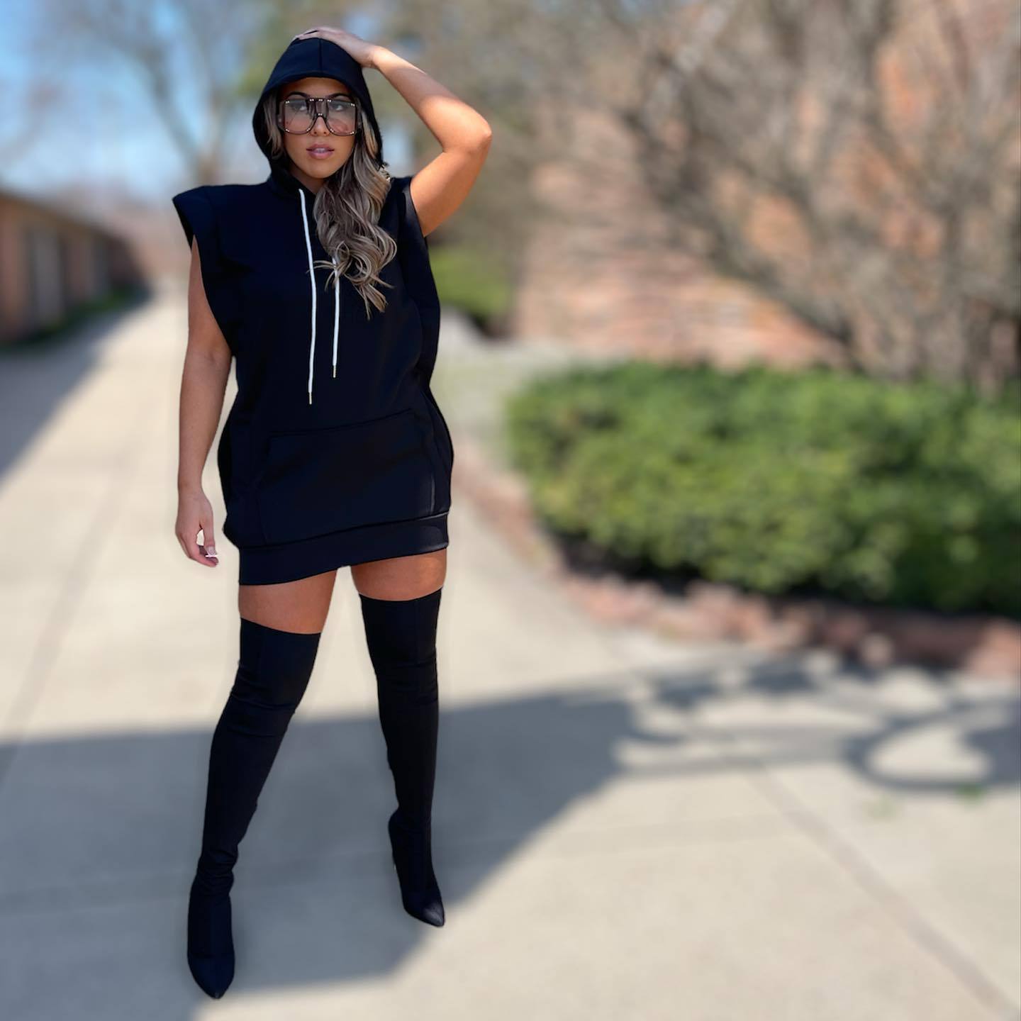 Woman wearing a black sleeveless hooded mini dress with white drawstrings and black thigh-high boots, posing outdoors.