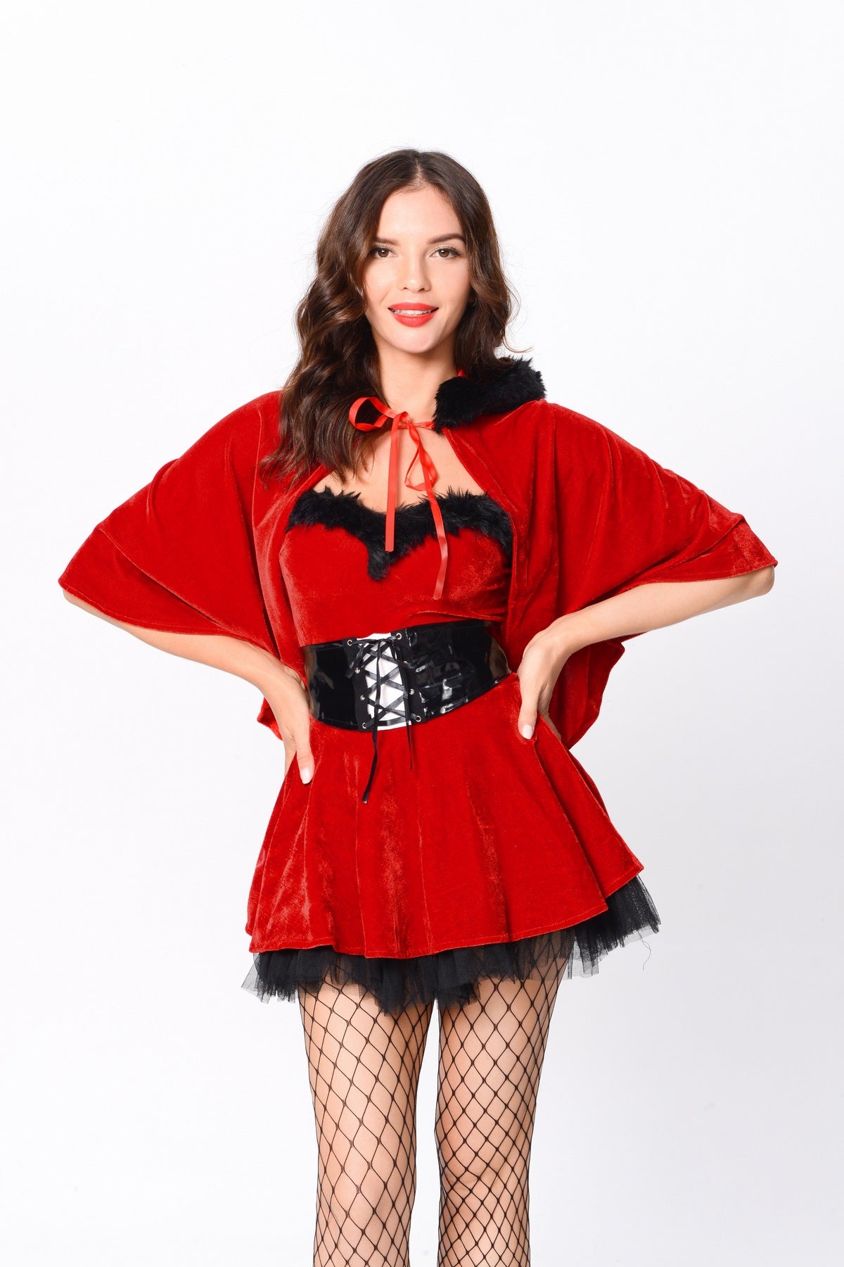 Woman wearing a red velvet costume with a cape, corset belt, and fishnet stockings, posing with hands on hips.
