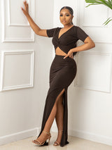 Woman wearing a dark brown two-piece set with a twisted v-neck top and a high slit skirt, posing with her hand on a white wall.