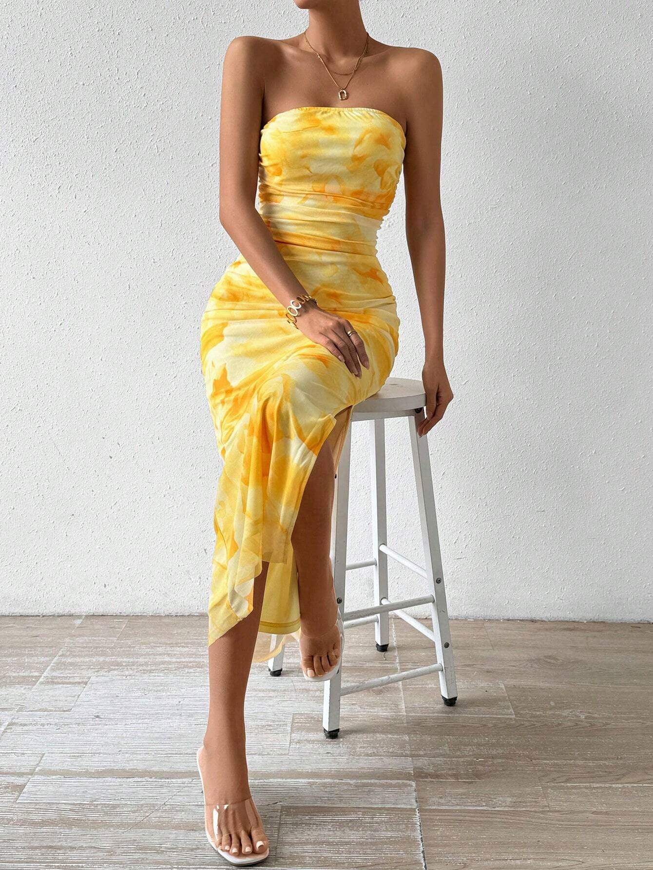 A woman sits on a white stool wearing a yellow tie-dye print strapless maxi dress.