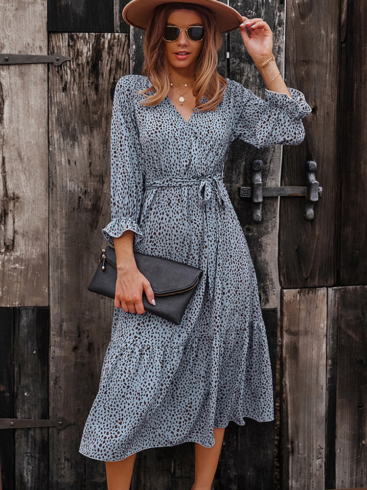 Woman wearing a light blue and brown polka dot midi dress, brown hat, sunglasses, and holding a black clutch.
