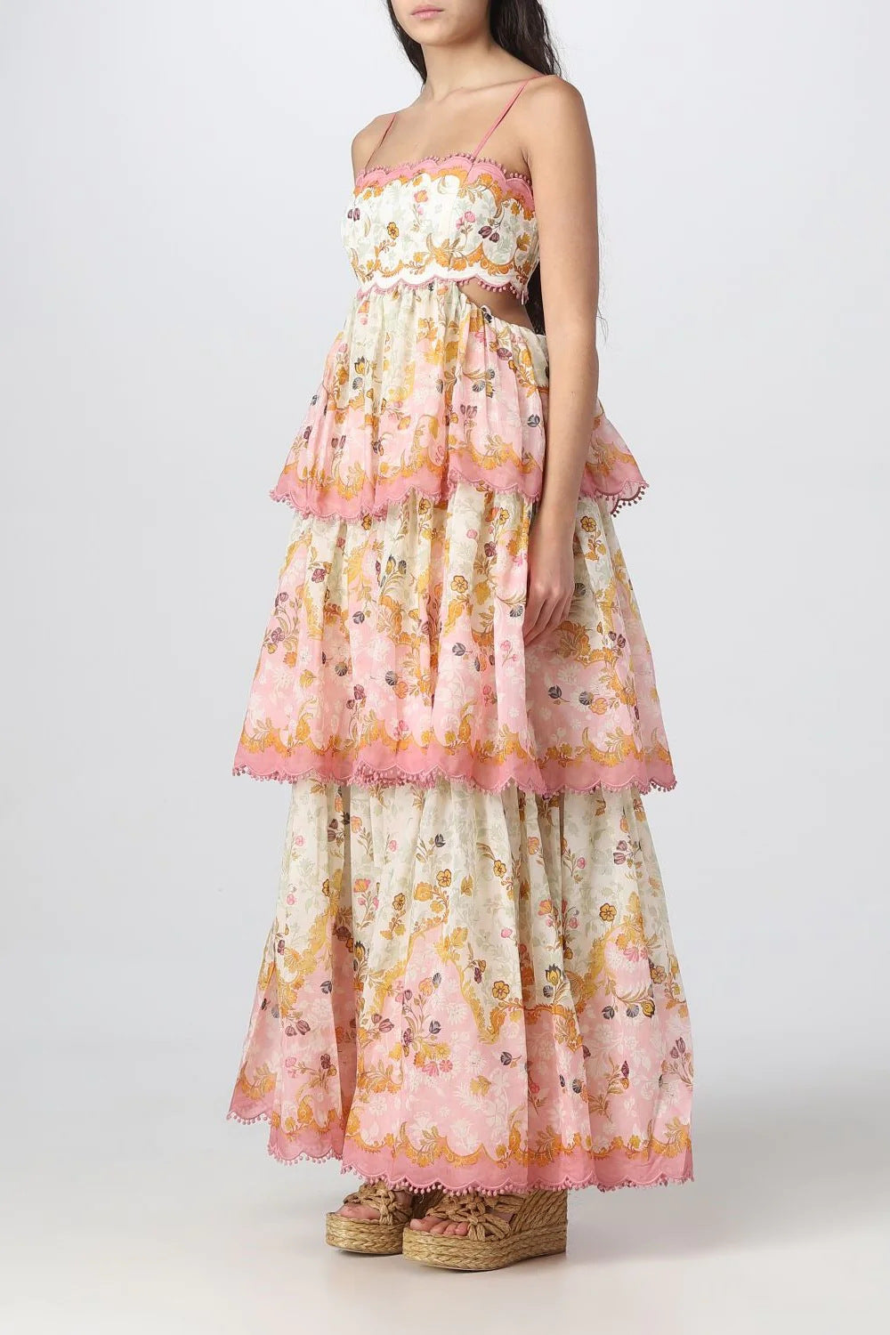 Side view of a woman wearing a tiered, floral print maxi dress with a scalloped and pom-pom trim.