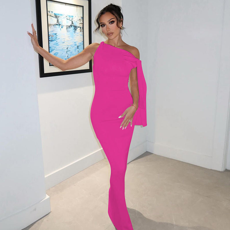 Woman in a hot pink sleeveless column gown with an asymmetrical neckline leaning against a white wall.