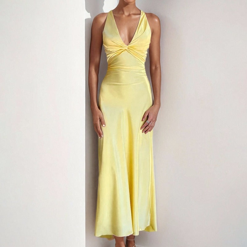 Woman wearing a yellow satin maxi dress with a twisted V-neckline and halter straps, leaning against a white wall.