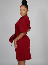 Side view of a woman in a red knee-length dress with puffed shoulders and a draped knot detail.