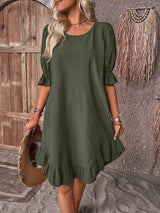Woman wearing a dark green short puff sleeve A-line dress with a ruffled hem and neckline, holding a straw hat.