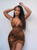 Woman with dark hair in a brown satin dress with a plunging neckline and high slit, posing against a white wall.