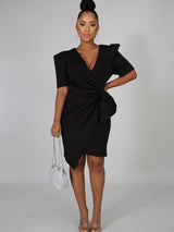 Woman wearing a black knee-length dress with puffed shoulders and a draped knot detail on the front.