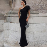 Woman in a black one-shoulder floor-length dress with feather shoulder detail and a silver clutch, standing near a stone building.
