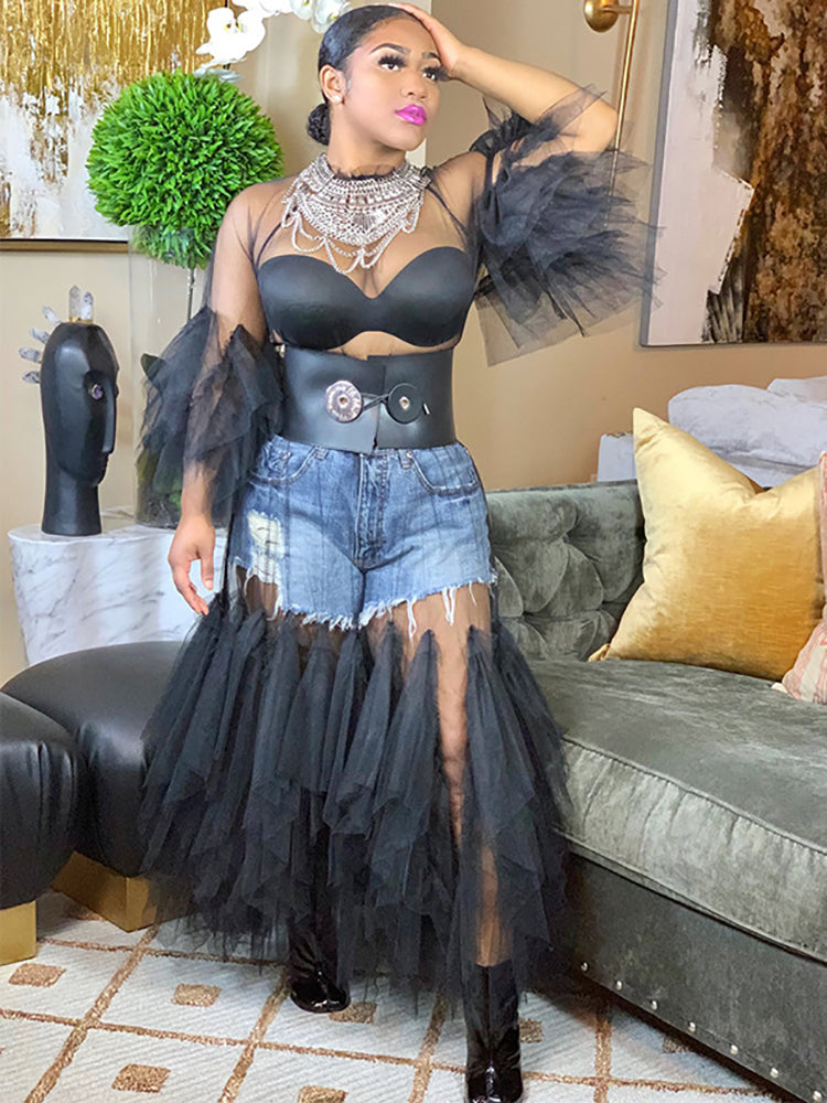 Woman wearing a black sheer mesh maxi dress with puffed sleeves and ruffle trim, paired with denim shorts, a black belt, and statement necklace.