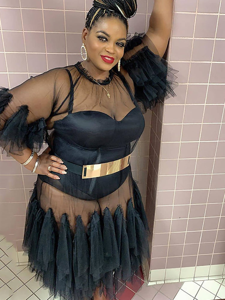 Close-up of a woman wearing a black sheer mesh bodysuit with a gold metallic belt, layered under a sheer black mesh dress with ruffle details.