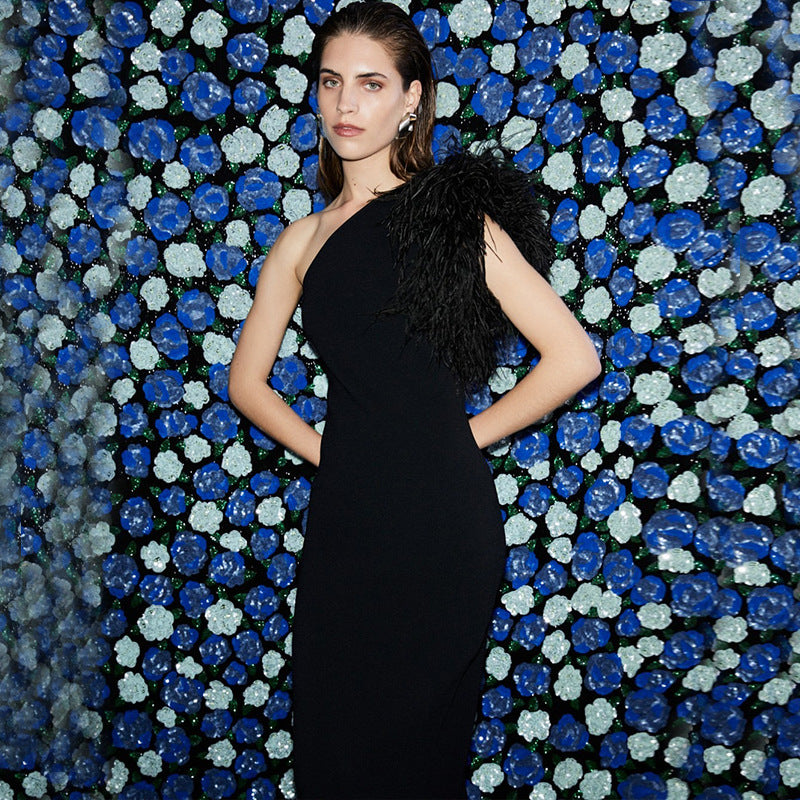 Woman in a black one-shoulder gown with feather accent posing against a blue and white floral backdrop.