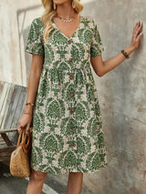 Woman leaning against a textured wall in a green and white floral print midi dress, holding a straw handbag.