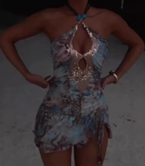Woman standing in a light blue and brown abstract print halter neck mini dress with a sequined keyhole neckline and turquoise necklace, with hands on her hips.