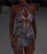 Woman walking forward in a light blue and brown abstract print halter neck mini dress with a sequined keyhole neckline and turquoise necklace.