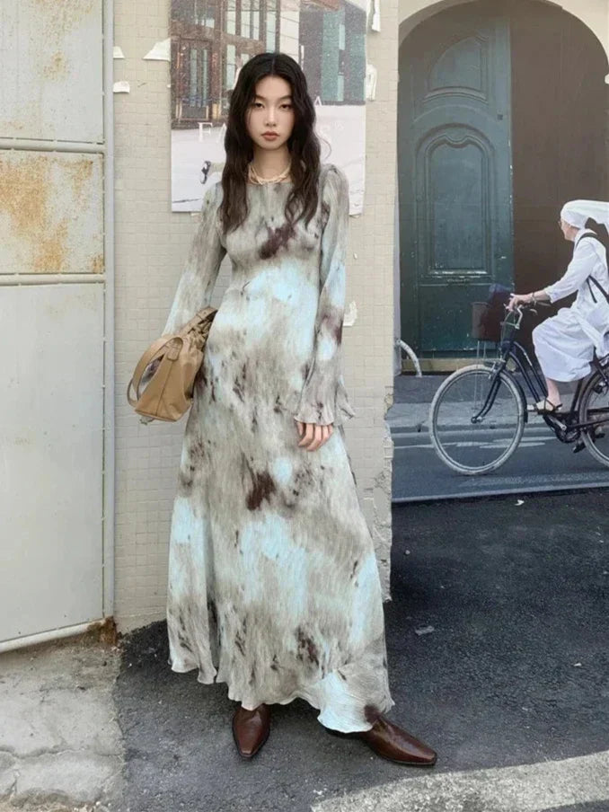 Woman wearing a long-sleeved, tie-dye maxi dress with brown boots and a tan handbag, standing outdoors.