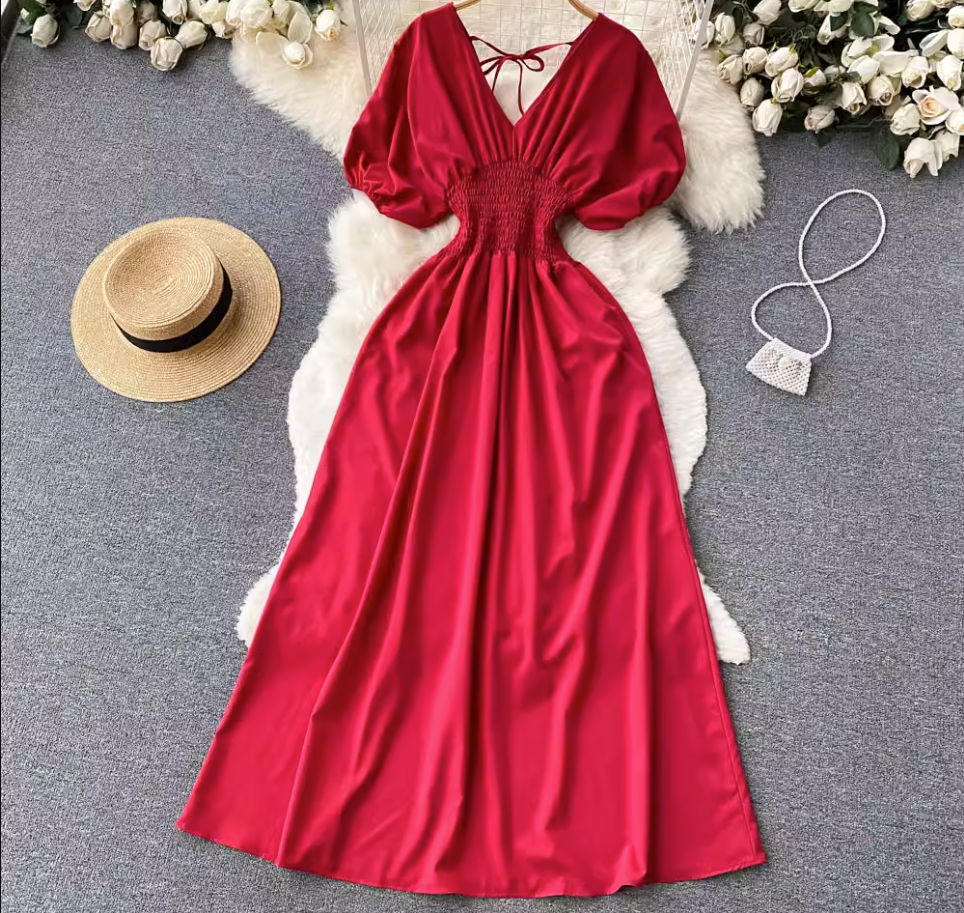Red maxi dress with smocked waist and puff sleeves laid flat on a fluffy rug with a straw hat and a small beaded purse