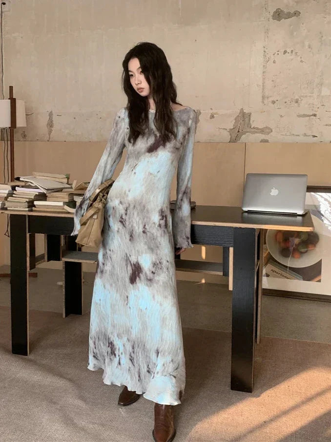 Woman in a tie-dye maxi dress posing indoors near a desk with books and a laptop.
