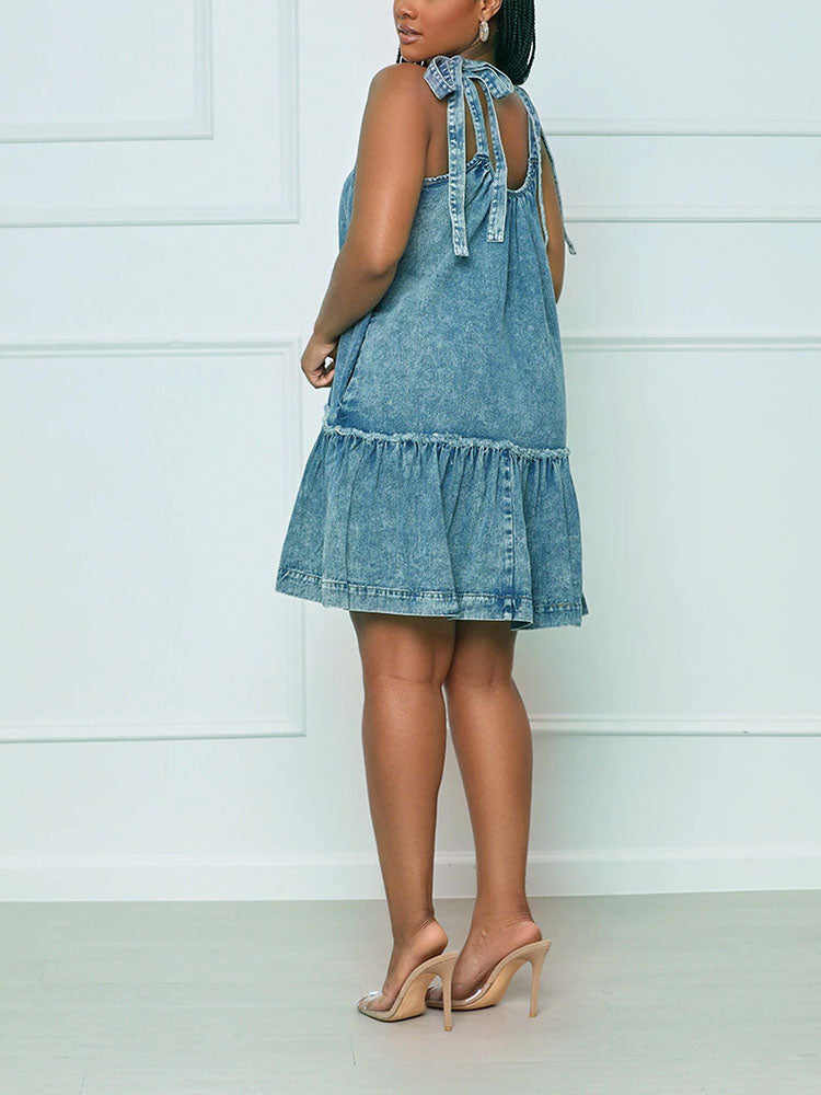 Woman wearing a blue denim A-line dress with tie shoulder straps and a ruffled hem, shown from the back.