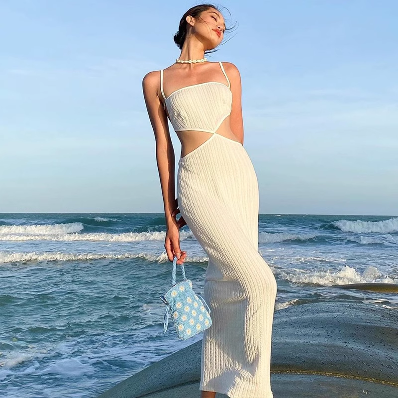 Woman wearing a white knit midi dress with cut-out details and a small blue bag, standing by the ocean.