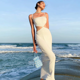 Woman wearing a white knit midi dress with cut-out details and a small blue bag, standing by the ocean.