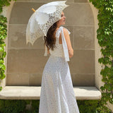 Back view of a woman wearing a white floral print maxi dress with tie shoulder straps and a high slit, holding a white lace parasol.