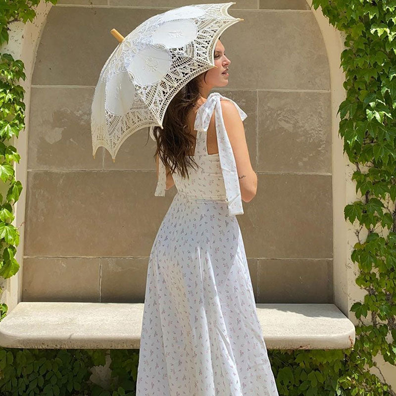 Back view of a woman wearing a white floral print maxi dress with tie shoulder straps and a high slit, holding a white lace parasol.