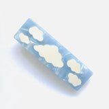 A rectangular barrette with a light blue marbled acetate base adorned with white cloud shapes.