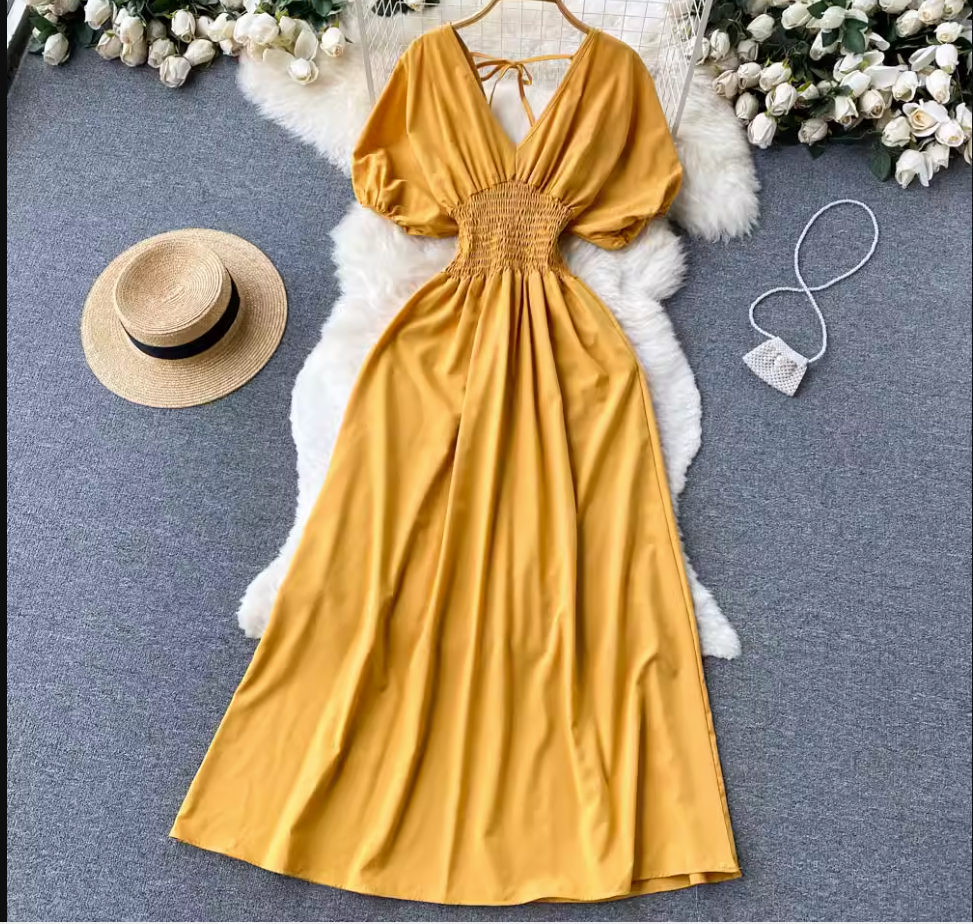 Yellow maxi dress with smocked waist and puff sleeves laid flat on a fluffy rug with a straw hat and a small beaded purse