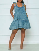 Woman wearing a blue denim A-line dress with tie shoulder straps and a ruffled hem, shown from the front with hands in pockets.