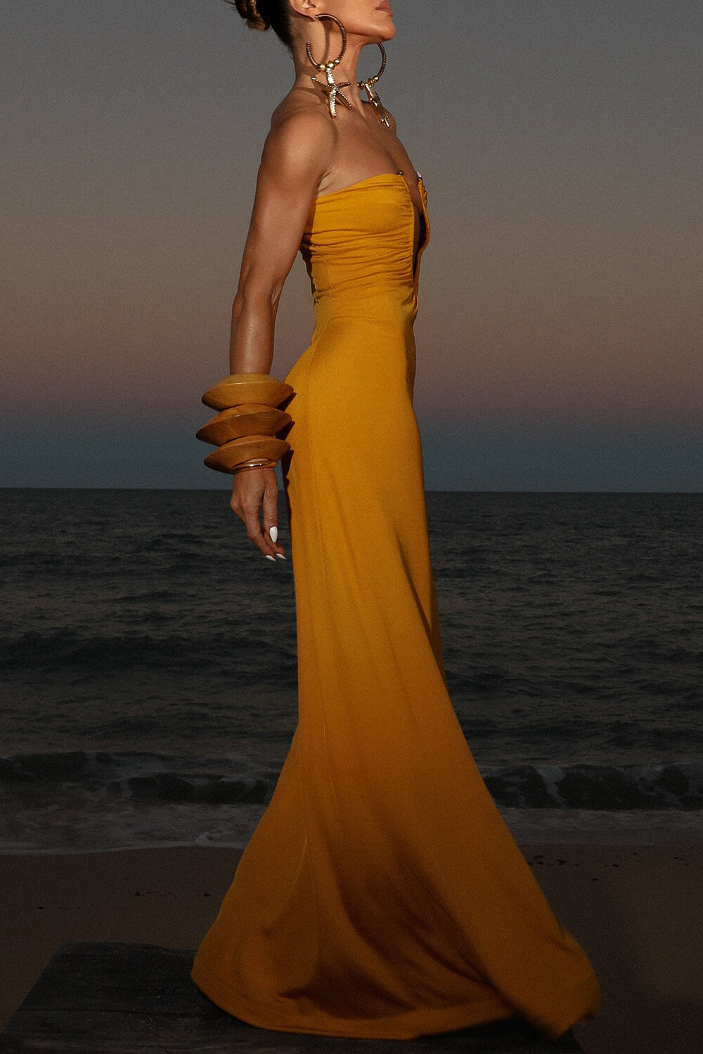 Side profile of a woman in a yellow floor-length gown with a deep V-neckline and ruched detailing, on a beach.