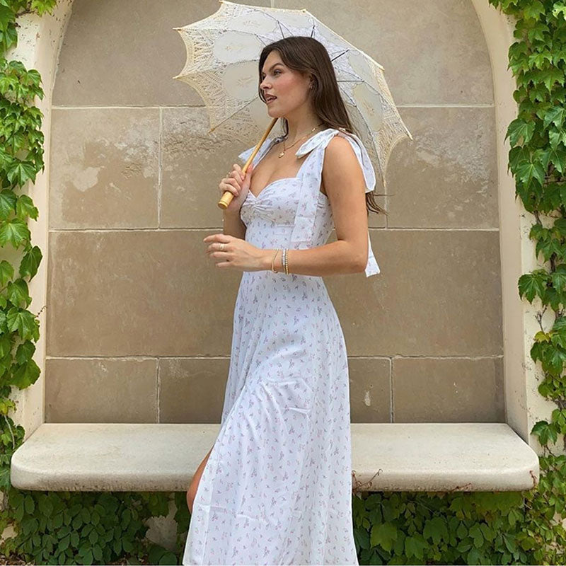 Woman wearing a white floral print maxi dress with tie shoulder straps and a high slit, holding a white lace parasol in front of a stone wall with ivy.