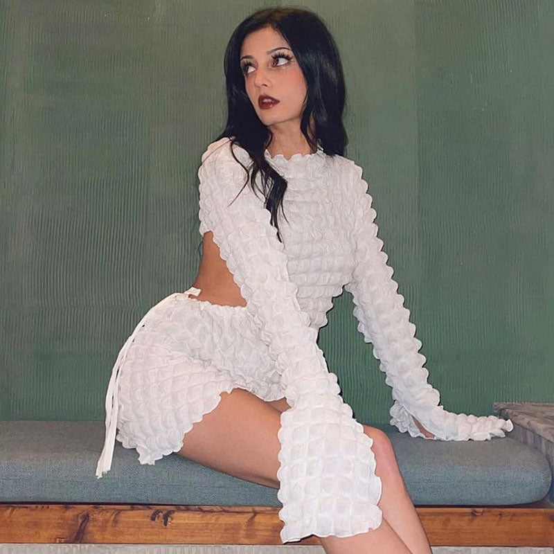 Woman in a white textured crinkle fabric mini dress with cut-out details and long bell sleeves, posing on a bench.