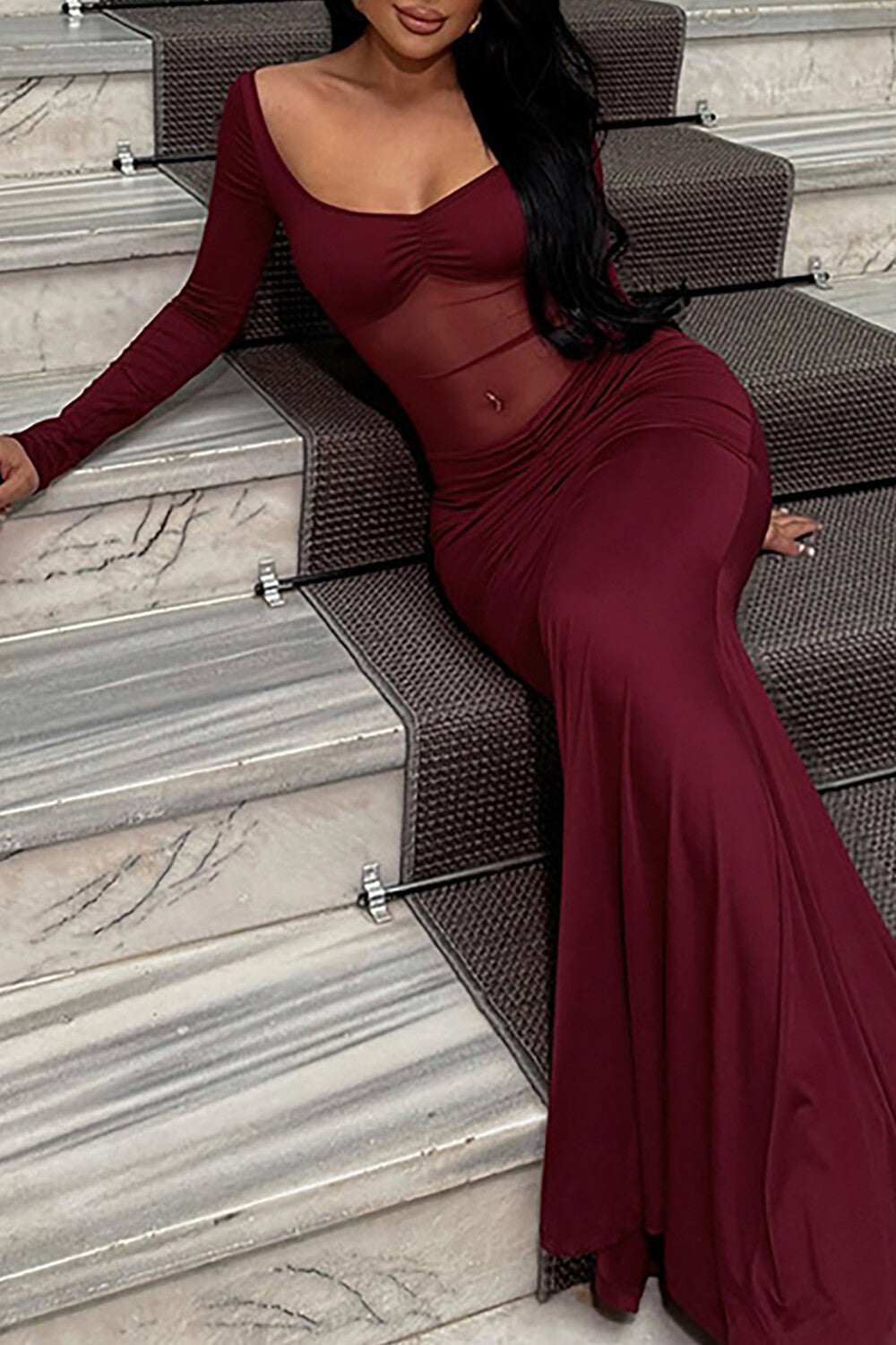 Woman in a burgundy long-sleeved mermaid dress with a sheer bodice and ruched waist, sitting on a staircase.