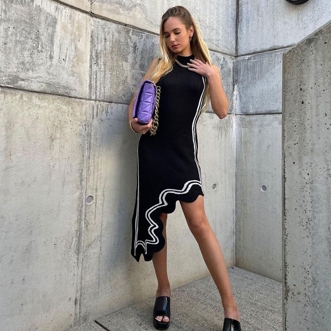 woman in black sleeveless dress with white piping and asymmetrical hem, holding a purple handbag