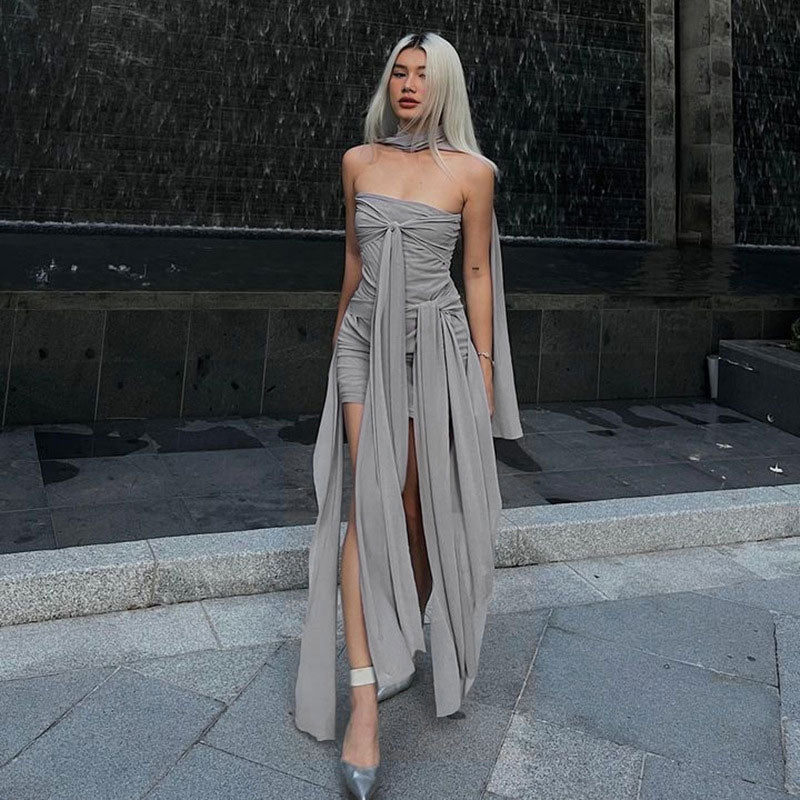 Woman wearing a grey strapless dress with flowing draped fabric and a high slit, standing in front of a water feature.