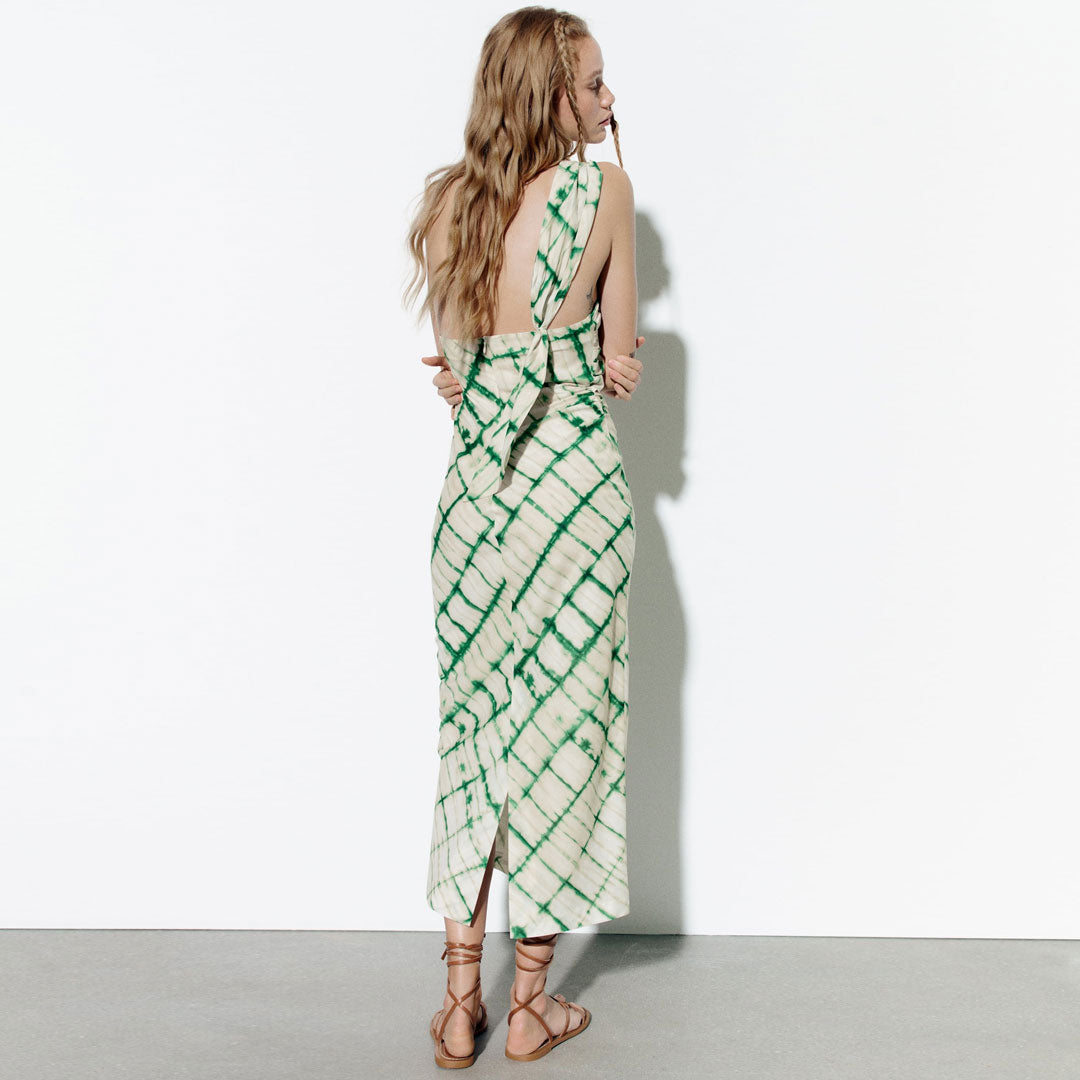 Back view of a woman in a green and cream tie-dye column dress with an asymmetrical halter neckline and a back tie detail.