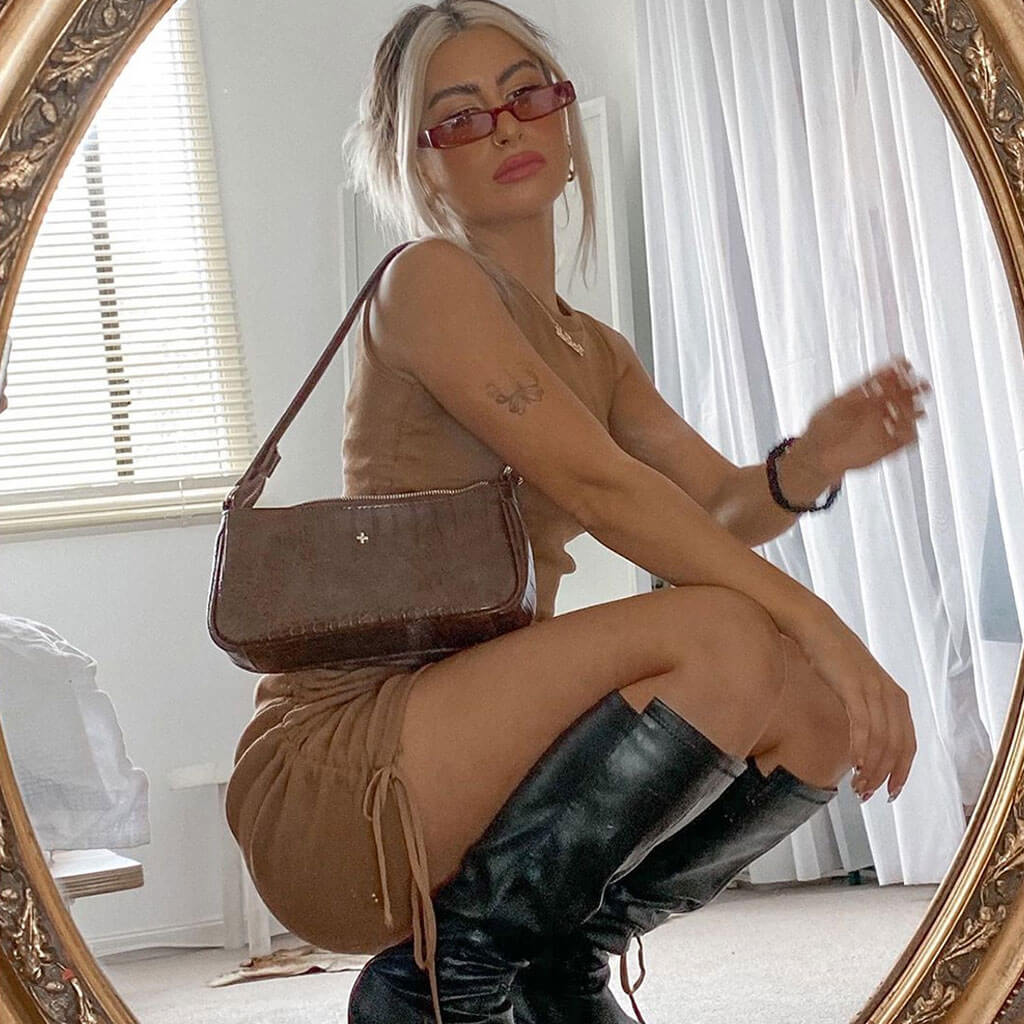 A woman wearing a brown ruched dress and black boots poses in front of a mirror.