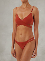 Woman wearing a rust-colored bikini with a crisscross halter top and double strap bottoms against a neutral background