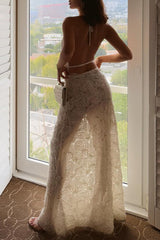Woman standing by a window wearing a white halter neck top and a sheer white maxi skirt with rose appliqué