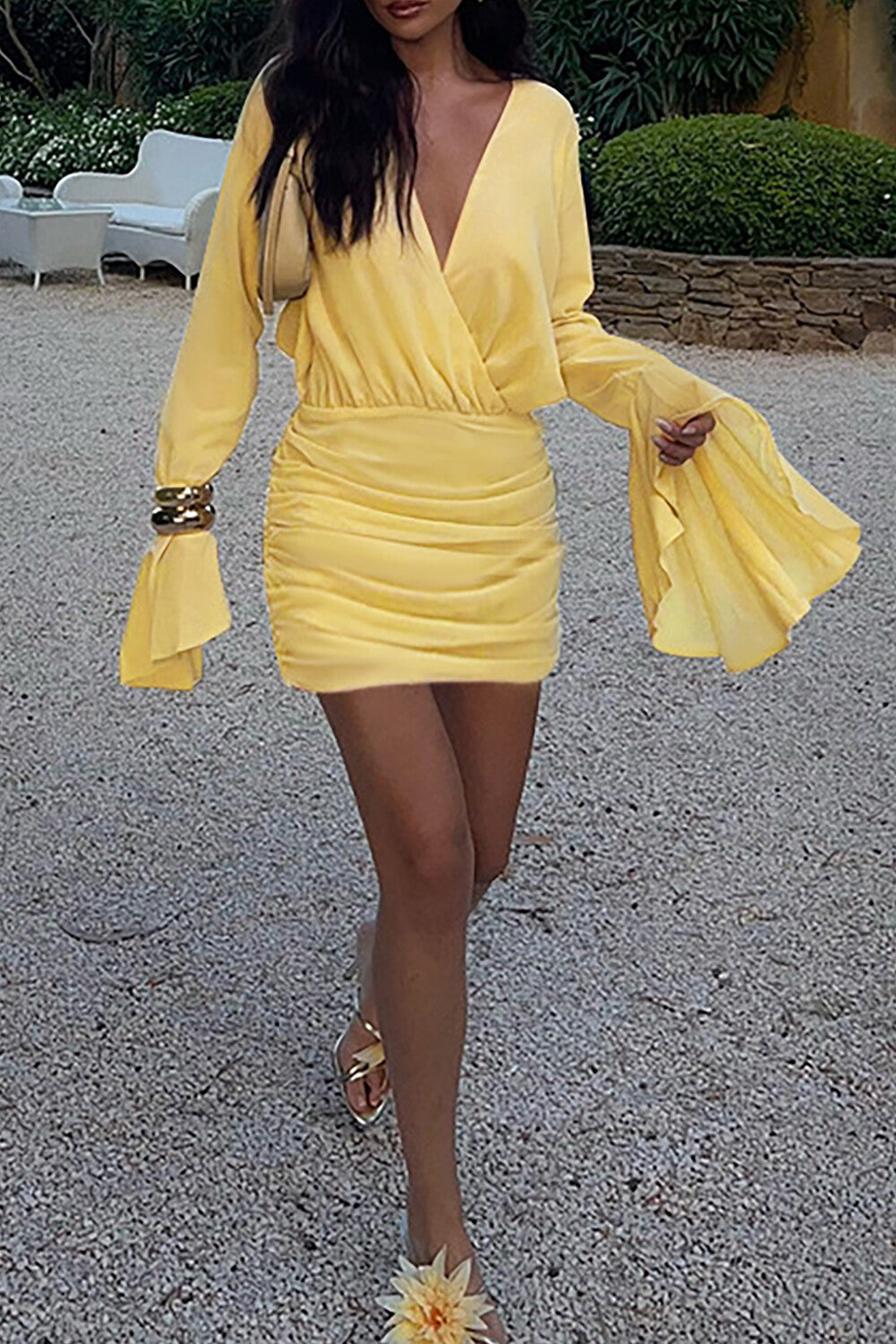 Woman walking in a yellow ruched mini dress with bell sleeves and gold sandals