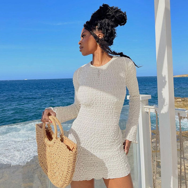 Woman in a white textured long-sleeve mini dress holding a straw bag, standing by the ocean.