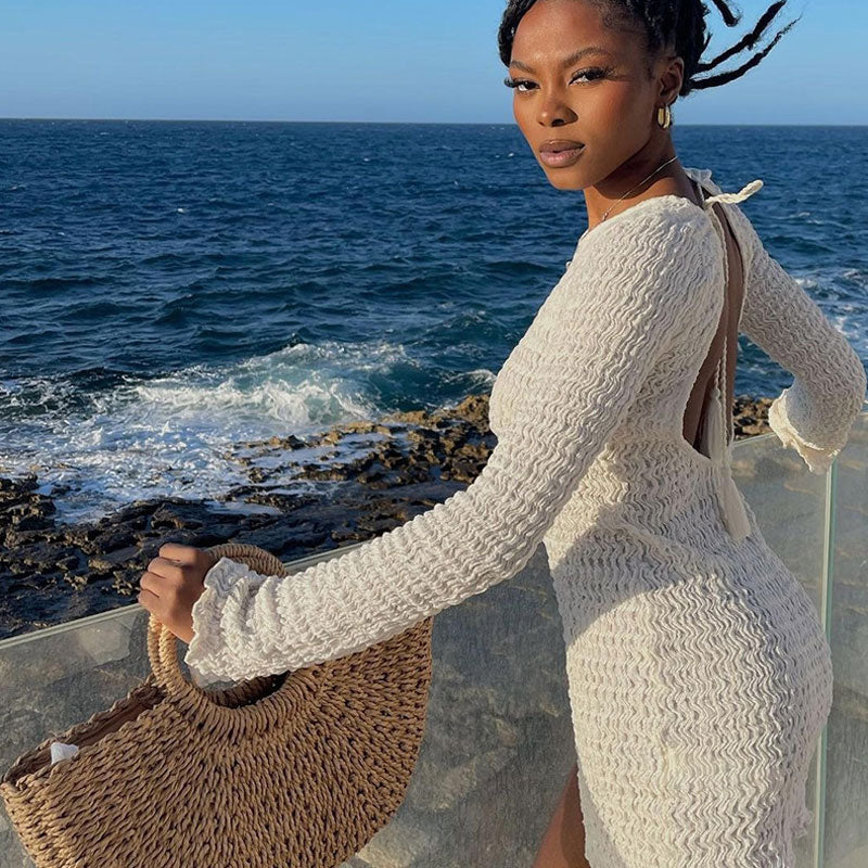 Woman in a white textured long-sleeve mini dress with an open back, standing by the ocean.